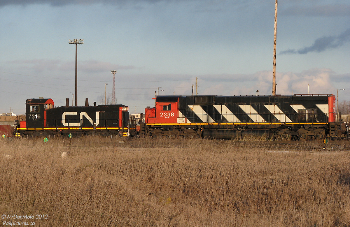 Across the field and far away. Sitting in the deadline behind the diesel shop, two retired relics of days past linger together on a November evening. CN M636 2338 once hauled heavy transcontinental freights with its 3600 horsepower engine roaring in notch 8. Retired in the late 1990's, she survived the scrapper as the backup Y2K power unit for the shops and has been here ever since, awaiting possible preservation or possible scrapping. SW1200RSm 7316, built in the 1950's for local and roadswitcher service, was upgraded and rebuilt by CN in the 80's, but eventually succumbed to a seized crankshaft that was then cut out. As of late she's been a parts unit for the nearby steel transfer facility's switcher RT-110.  Fast forward a few years: 2338 survived on long enough to be purchased by the WNY&P and transported to the US as RPRX 2338, although CN held her up from moving suspiciously long enough for someone to get a photo with the "new" CN 2338 (GE ES44DC) posed side-by-side (methinks there was a railfan in the diesel shop that held it up until then...). 7316 was sold to LDS in Sarnia, where their capable shop will evaluate her for possible rebuild and resale.  Both have eluded the scrapper's torch, for now.