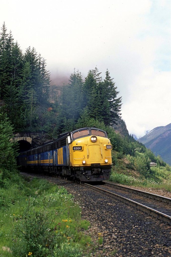 The head end of the eastbound "Canadian" roars out of the small tunnel near the west switch of Cathedral in Kicking Horse Pass. It is a long train this day. The rear car can be seen at right on the ledge that the train has been ascending on since leaving Field.