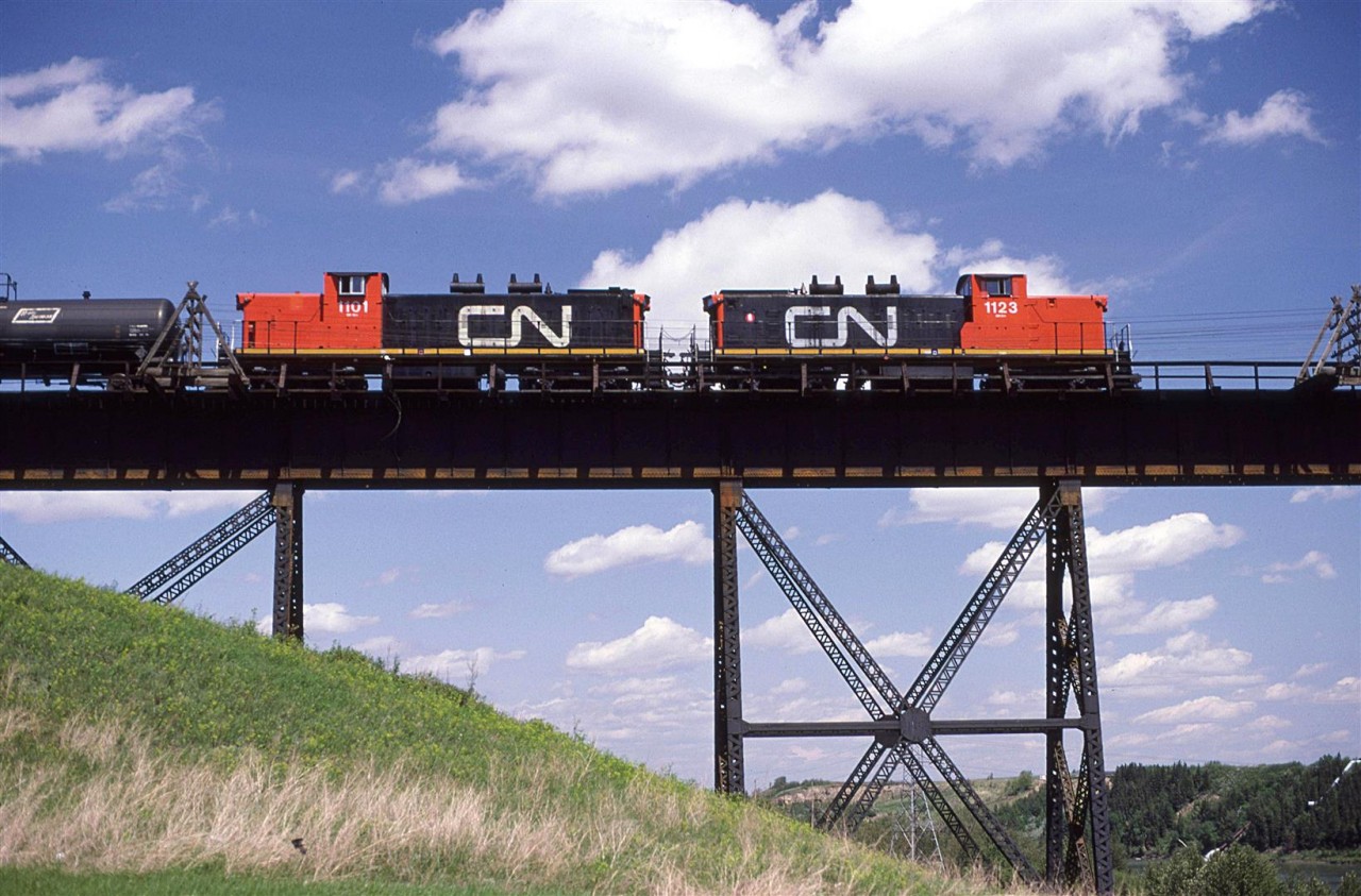 This is likely, but not necessarily an eastbound Clover Bar Transfer (Calder to Clover Bar Yard) beginning the crossing of the North Saskatchewan River in NE Edmonton.
The units are re-trucked GMD-1's. They were to become very common for switch in transfer duties in Edmonton. The need for the 1A1 trucks in the Edmonton area (and likely system wide) was diminishing at this time.
Those telegraph cable outriggers on the bridge would be removed sometime around 1991 when CN decides to rid the landscape of the poles and cables.