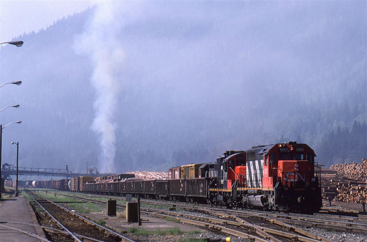 On a very calm summer morning in Boston Bar, the sky fills with smoke from the lumber yard. A couple CN manifest trains await a crew for their eastward journey.
I think that everything associated with the mill is gone.