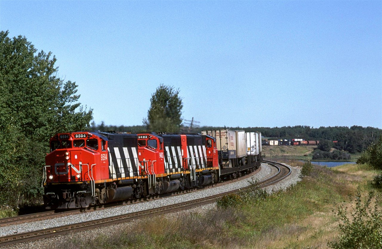 This is a westbound train swinging through the big S curve at Carvel. Mink Lake is to the right. An eastbound train, perhaps 358/360 with a few empty grain cars stuck on the end, is occupying the south track.