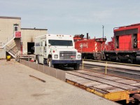 Flat car with a Brinks truck load by the materials department with SW1200rs hump unit 1159 in the background.