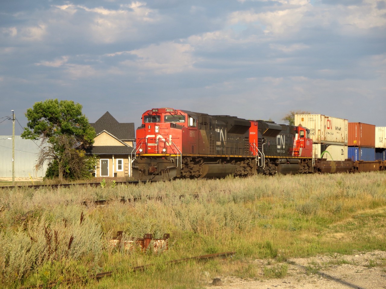 CN 8816 West rolls through Elie, MB with a 107 platform intermodal train as evening approaches on the CN Rivers Sub. Within a mintute they were met by CN B730 with CN 3117 leading 204 Eastbound potash loads. 
In the background is the former Canadian Northern station, which has been moved off its original site to a spot a little farther from the tracks. It has been fixed up and now is someone's residence.