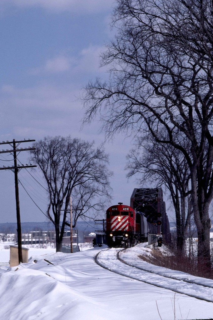 The Nackawic Turn is close to its home terminal in Fredericton, New Brunswick, on a beautiful afternoon in March 1985.  It is about to pass the site of the former CNR Fredericton station on the engineer's side of the tracks.  

C-424 4218 was built in Montreal, Quebec, by MLW and delivered as the 8318 on Tuesday, October 5, 1965, with serial 84856.  The CPR received trade-in credit with the return of Alco FB-1 4402 on Thursday, July 8.  The unit was part of 32 unit order 4905, class DRF-24b, built between March and December 1965.  Their delivery was followed immediately by order 4906 for 18 units of class DRF-24c.  MLW built these without trade-ins from December 1965 through March 1966.  CPR initially used the C-424s on priority freights, notably on the Eastern Region, as St. Luc was their original maintenance assignment.  When they were new, I often saw them on Windsor - Montreal hotshots in combination with the nearly new GP35s, which had proven slippery.  CPR quickly renumbered the units in the 4200 series.  Retirement, conversion or sale came to the C-424s before 1996; however, the 4218 was not one of the 31 survivors.  

The Nackawic Turn left Fredericton weekday mornings with traffic for the paper mill at Nackawic on the north side of the Saint John River.  At Una Junction, Mileage 21.8 of the Fredericton Sub, the outbound train turned north on trackage rights at Mileage 0.2 of the CNR Fredericton Spur and joined their 70.5-mile Saint John - South Devon Oromocto Sub at Mileage 69.4.  The Turn then crossed the bridge in the background.  The ice blocked river destroyed the original crossing; this bridge opened in 1936.  (It survives as a walking path following the end of rail service in the late 1990s.)   Reaching South Devon on the northern bank, the train turned north for a 37-mile run to Southampton on the Gibson Sub.  At Southampton, Mileage 22, it turned south, travelling 9.4 miles to the mill at Nackawic.   

Upon returning to Fredericton, the power was set up to do a nightly 133.2-mile turn to Saint John.  This arrangement ended on Friday, October 25, 1985, thereafter power from mainline freights brought cars the 22.2 miles into town from Fredericton Junction on the Montreal - Saint John line.   Prior to Friday, November 28, 1980, the nightly freight, the "Oriental', took its train to McAdam, forty miles west of the Junction.  

The 1967 flooding behind the Mactaquac dam caused the relocation of several communities. It spawned the province's first model community in Nackawic.  The flooding also cut short the last 3.1 miles of the Southampton Sub that reached down to the Saint John River at Otis.  St. Anne Nackawic Pulp and Paper opened their kraft pulping mill in 1970 and received a continuing supply of rail-hauled Bunker 'C' oil from the Irving refinery in Saint John.   Rail shipments lasted until December 1994 when CP Rail discontinued its operations east of St. Jean, Quebec and abandoned its branchlines north of Fredericton.   The present owner, AV Group NB Inc., produces up to 540 metric tons of bleach kraft pulp daily.