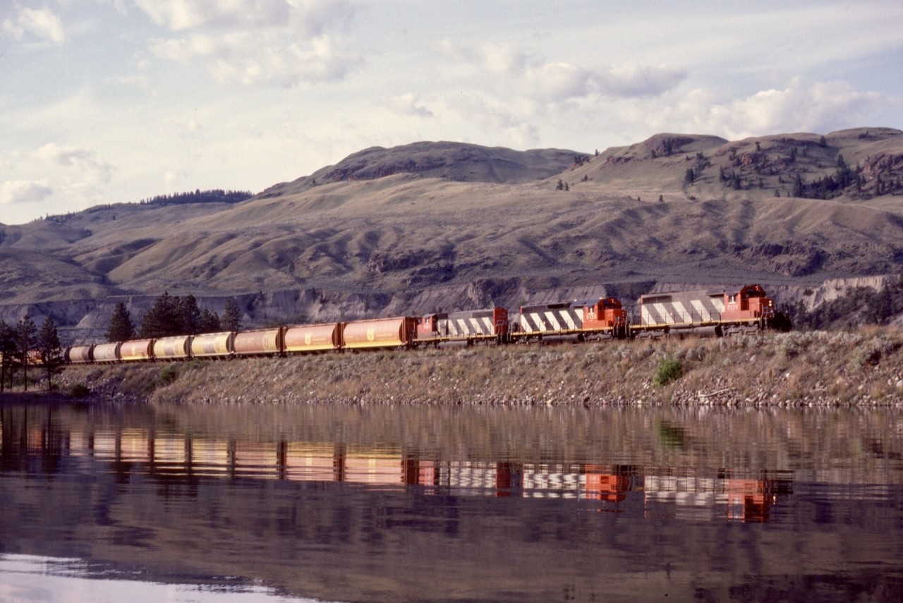 West from Kamloops, CN stays fairly close to water level, first along the north side of Kamloops Lake then along the Thompson River.  Shortly before reaching Walhachin, an old river oxbow is crossed, and early morning near-summer lighting is ideal for catching reflections, illustrated here at 0708 PDT on Tuesday 1982-06-01 with a grain train westward behind CN 5139 + 5125 + 5280 at mileage 29.9.

The cut face of the distant bank is for CP’s Thompson sub. ascending to a summit at Walhachin.