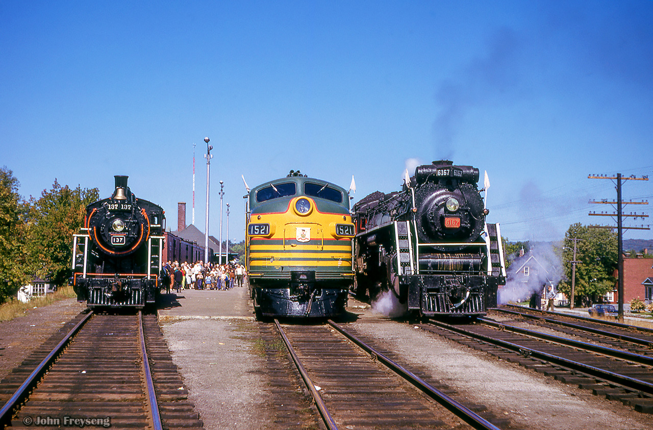 A lineup of power at North Bay greets photographers aboard the Upper Canada Railway Society's massive undertaking of an excursion: a 692.2 mile trip from Toronto - Ottawa - North Bay - Temagami - Toronto, with seventeen coaches behind CNR 6167.  With the train having run overnight from Toronto - Ottawa on the 13th, the train turned west and departed the nation's capital early on the 14th. 6167 would be removed at North Bay, with ONR FP7s 1521 and 1511 handling the train to Temagami overnight.

Here, the consist has just returned from Temagami and a change of engines will be taking place shortly.  For now, photographers scramble to the head end to capture the moment.  At left is Temiskaming & Northern Ontario Railway 2-8-0 137, originally built in 1913 as Canadian Northern Railway M-3-e class 2164, and later to the Canadian National with the same number.  The engine was sold to the Ontario Northland Railway in February 1963, and displayed at Cobalt during the first two weeks of August as part of the town's 60th anniversary celebrations.  T&NO 137 later operated in excursion service during 1967 as part of Canada's centennial celebrations, and later at different times until final retirement in 1972.  It is now on display at Cochrane.  Front and centre is ONR FP7s 1521 and 1511, built by GMDD London in 1953 and 1952 respectively.  1521 would be rebuilt to ONT 2002 in 1997, retired in 2004, and put on "display" in North Bay, though it still sits at the north end of ONR's yard near their head office.  Trailing unit 1511 would be sold to GO Transit, becoming their APCU 906, and later scrapped in 1995.  At right, CNR 6167 stands ready to lead the train home to Toronto.

Today this scene still looks similar, minus the tracks.  The buildings at right still stand on 3rd Avenue, as does the station and empty platforms, built in 1916 by the Canadian Northern.  Much of the CNR right of way through North Bay has vanished as this portion of the Alderdale Sub from Nipissing to Capreol was reassigned as an extension of the Newmarket Sub in 1988.  CN applied to abandon the Newmarket from Yellek (6 miles west of North Bay) to Capreol in 1996, with removal by CANDO to follow in 1997.  



Scan and editing by Jacob Patterson.