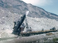 A rare view of a Canadian Pacific rail-mounted steam shovel, CP 400308, loading some ballast flatcars with loads of rock at Frank Slide, Alberta. A worker rides the deck behind the shovel boom, likely operating the controls for its large chain-driven shovel bucket, as more workers set up the trailing flatcars with wooden stakes and blocking. The steam shovel is operating in the middle of the namesake rockslide, on some siding tracks off CP's Crowsnest Subdivision mainline. Information on CP's operation here is scarce, the shovel might have been stationed here for conveniently obtaining aggregate from the slide for railway purposes (fill, crushing, etc). Rock loading on flatcars with wooden stakes and side rails is also rather curious, although due to the size of the rocks, removing the stakes and pushing them off the sides may have been the easiest way to unload them (they would have clogged bottom-dump hopper cars, and would have been harder to unload from gondolas).<br><br>Frank Slide got its name on April 29th 1903 when a giant rockslide of limestone slid down Turtle Mountain and buried the east end of the mining town of Frank, the coal mine, the CPR line, and buried between 70-90 residents of the town. The railway line and mine were both opened, part of the town relocated further away, but much of the area remains unchanged since the rockslide happened.<br><br>With the exception of some early images, photos of CP steam shovels in their later years are scarce. An old freight car roster shows three steam shovels (CP 400308-400310, each with a 4 cubic yard bucket) still on the roster in 1965, although with the popularity of newer, more nimble diesel-powered cable and hydraulic excavators, those old steam-powered relics likely didn't last too much longer.<br><br><i>Original photographer unknown (possibly E.W.Johnson of Calgary), Dan Dell'Unto collection slide with extensive restoration (this was the first slide in a roll, and 1/3rd of it was horribly overexposed and colour-shifted).