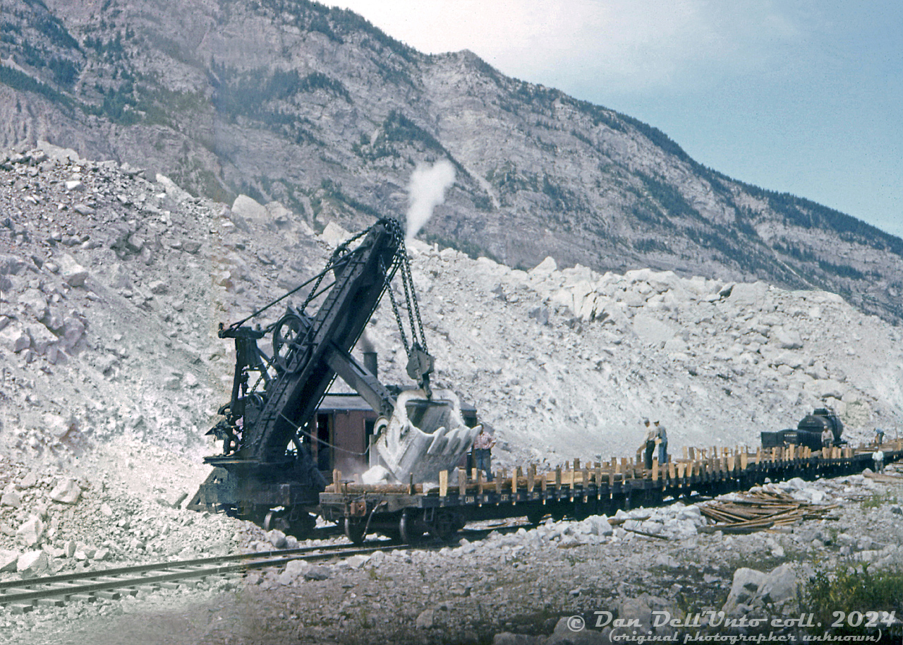 A rare view of a Canadian Pacific rail-mounted steam shovel, CP 400308, loading some ballast flatcars with loads of rock at Frank Slide, Alberta. A worker rides the deck behind the shovel boom, likely operating the controls for its large chain-driven shovel bucket, as more workers set up the trailing flatcars with wooden stakes and blocking. The steam shovel is operating in the middle of the namesake rockslide, on some siding tracks off CP's Crowsnest Subdivision mainline. Information on CP's operation here is scarce, the shovel might have been stationed here for conveniently obtaining aggregate from the slide for railway purposes (fill, crushing, etc). Rock loading on flatcars with wooden stakes and side rails is also rather curious, although due to the size of the rocks, removing the stakes and pushing them off the sides may have been the easiest way to unload them (they would have clogged bottom-dump hopper cars, and would have been harder to unload from gondolas).Frank Slide got its name on April 29th 1903 when a giant rockslide of limestone slid down Turtle Mountain and buried the east end of the mining town of Frank, the coal mine, the CPR line, and buried between 70-90 residents of the town. The railway line and mine were both opened, part of the town relocated further away, but much of the area remains unchanged since the rockslide happened.With the exception of some early images, photos of CP steam shovels in their later years are scarce. An old freight car roster shows three steam shovels (CP 400308-400310, each with a 4 cubic yard bucket) still on the roster in 1965, although with the popularity of newer, more nimble diesel-powered cable and hydraulic excavators, those old steam-powered relics likely didn't last too much longer.Original photographer unknown (possibly E.W.Johnson of Calgary), Dan Dell'Unto collection slide with extensive restoration (this was the first slide in a roll, and 1/3rd of it was horribly overexposed and colour-shifted).