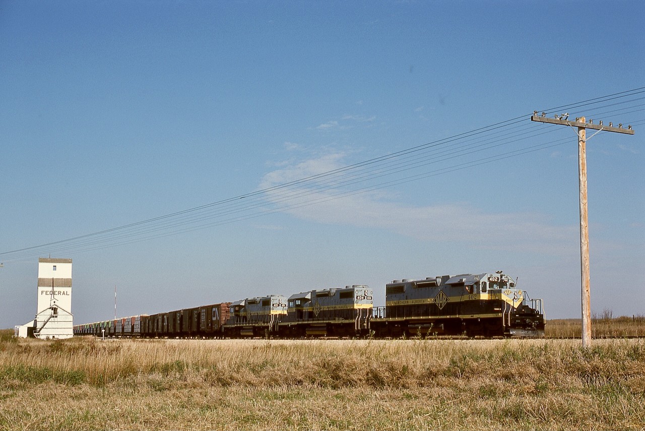 Northern Alberta Railways typically utilized three of their four SD38-2 units to handle the Great Slave Lake traffic down to and then up from a bridge across the Peace River at the town of the same name.  Here, on Friday 1976-09-24, that day’s GSL Turn is just starting to roll homeward again behind 402 + 404 + 401 after pausing for a mandatory standing train inspection after a hard grind upgrade from Peace River to level land at Judah, and now about to cross Township Road 825, with forty-one miles to Winagami Junction then one more to home terminal McLennan.  From there, two more subdivisions spread over 262 miles for the cars to reach Edmonton.