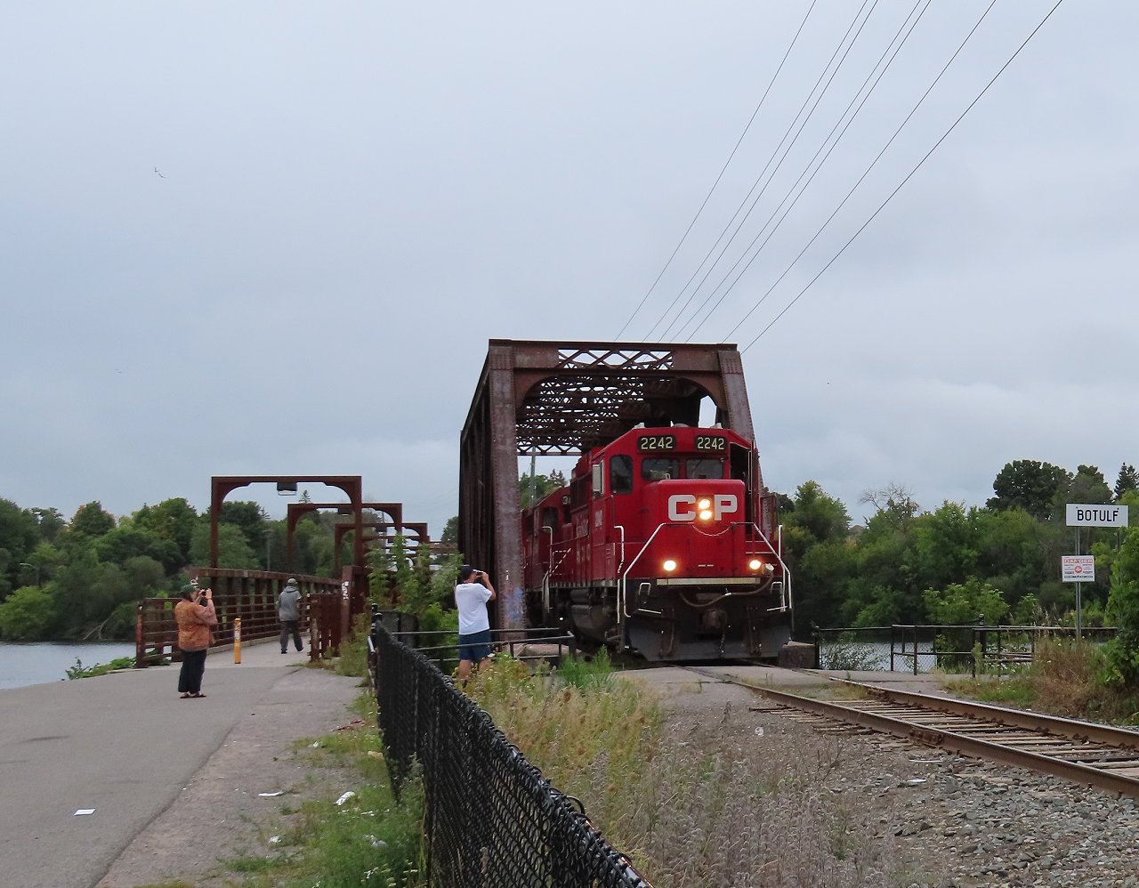 On a misty day, a couple of locals grab photos of the Havelock train as it exits the bridge over the Otonabee River. The pedestrian bridge to the left replaced an earlier narrow walkway that was cantilevered off the CP bridge.