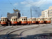 Toronto Transit Commission 2546, 2950, 2920, 2972, 2974, and 2958 are just some of the early 1920's Peter Witt streetcars stored in the TTC's temporary Harbour Yard on the harbourfront in downtown Toronto. These particular cars were "trailer train" consists with a "Large Witt" that hauled an unpowered trailer Witt, the extra capacity necessary for operation on the busy Yonge route between downtown and Glen Echo Loop.<br><br>Storage here was necessary due to construction of the new Yonge subway line's terminal facilities at Eglinton & Yonge, that interfering with streetcar storage and operation at the TTC's existing Eglinton Carhouse. The lack of space at Eglinton forced the TTC to store and operate its Yonge streetcars from this temporary 6-track yard, built on Toronto Harbour Commission lands at the south end of the busy Yonge streetcar route.<br><br>This lasted a few years (1951-1954) until subway construction was finished and the Yonge streetcar route was replaced with the new Yonge subway line (opened in March of the next year). Overnight, a lot of the old Peter Witt fleet suddenly became surplus, and they were rounded up and stored at Harbour Yard awaiting disposal for scrap or resale, including all of the trailers. After the Witts were cleared out, the temporary yard was removed. In the early 60's, streetcar tracks would return for a few years when <a href=http://www.railpictures.ca/?attachment_id=19956><b>Ferry Loop</b></a> was relocated.<br><br>Newer more modern PCC streetcars handled most of Toronto's streetcar route demands going forward, but a handful of Peter Witt streetcars remained in service until the <a href=http://www.railpictures.ca/?attachment_id=25110><b>University subway line opened in 1963</b></a>, and a final few saw limited use but were kept around until <a href=http://www.railpictures.ca/?attachment_id=47439><b>retirement in 1965</b></a>.<br><br>The temporary TTC Harbour Yard was located on the parcel of THC land bordered by Harbour Street, Bay Street, York Street and Queen's Quay. Yardley's Perfumes is visible in the background along York (it had a rail siding from nearby <a href=http://www.railpictures.ca/?attachment_id=54744><b>Rees St. Yard</b></a>). On the right is the brand new Workmen's Compensation Board Building at 90 Harbour Street (built 1953, demolished 2011). Downtown condos and modern office towers (and "Love Park") reside on the land here today.<br><br><i>Robert W. Gibson photo, Dan Dell'Unto collecton slide.</i>