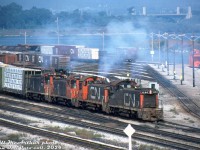 Things are looking more than a little hazy around CN's Stuart Street yard in Hamilton, as four GMD SW1200RS units (CN 1250, 1315, 1249 and 1254) on an eastbound freight coat the yard in a blue-white haze of diesel exhaust as they switch cars. 
<br><br>
Things of interest in this scene include the BC Rail bulkhead flatcars, CN 40' boxcars in the yard, a snowplow on the track to the side, and the high level York Blvd bridge in the background by Hamilton Junction.
<br><br>
<i>Bill McArthur photo, Dan Dell'Unto collection negative.</i>