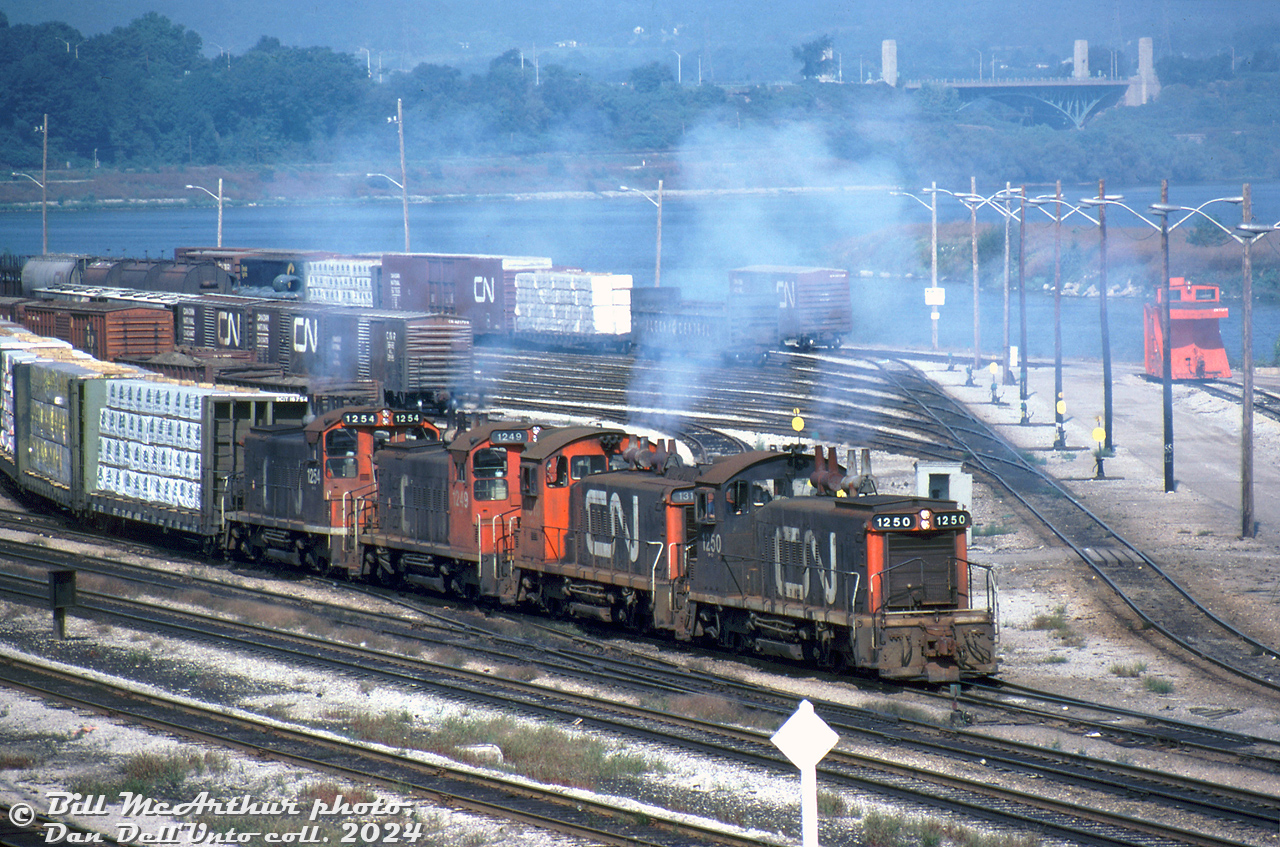 Things are looking more than a little hazy around CN's Stuart Street yard in Hamilton, as four GMD SW1200RS units (CN 1250, 1315, 1249 and 1254) on an eastbound freight coat the yard in a blue-white haze of diesel exhaust as they switch cars. 

Things of interest in this scene include the BC Rail bulkhead flatcars, CN 40' boxcars in the yard, a snowplow on the track to the side, and the high level York Blvd bridge in the background by Hamilton Junction.

Bill McArthur photo, Dan Dell'Unto collection negative.