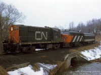 CN RS18 3118, FPA4 6779 and a single passenger car work their way back to the station on the Brockville Loop Line after visiting the Brockville wye (located amid a residential area to the southwest of the CN station area). After some research and input from others train appears to be heading eastbound on the north end of the Brockville Loop Line, crossing over a culvert for Butler's Creek and approaching Perth Street crossing. The houses in the background appear to be along Hubbell Street.<br><br>The wye trackage was part of the old Brockville, Westport & North-Western Railway, that ran between Brockville and Westport, Ontario. The line was acquired by Canadian Northern, and became part of Canadian National, who abandoned it in 1952. The wye and CN connecting track (the "Brockville Loop Line" that ran from the CN station area west, ducking under the Kingston Sub to get to the wye and BW&NW) was left intact, and used by CN and later VIA to wye passenger power and equipment as needed into at least the mid-80's. The wye and line is now part of the Brock Trail.<br><br>The processing date on this slide was stamped June 1973, but judging by the snow and sparse foliage, this may be a March or April photo.<br><br><i>Al Gorney photo, Dan Dell'Unto collection slide.</i>