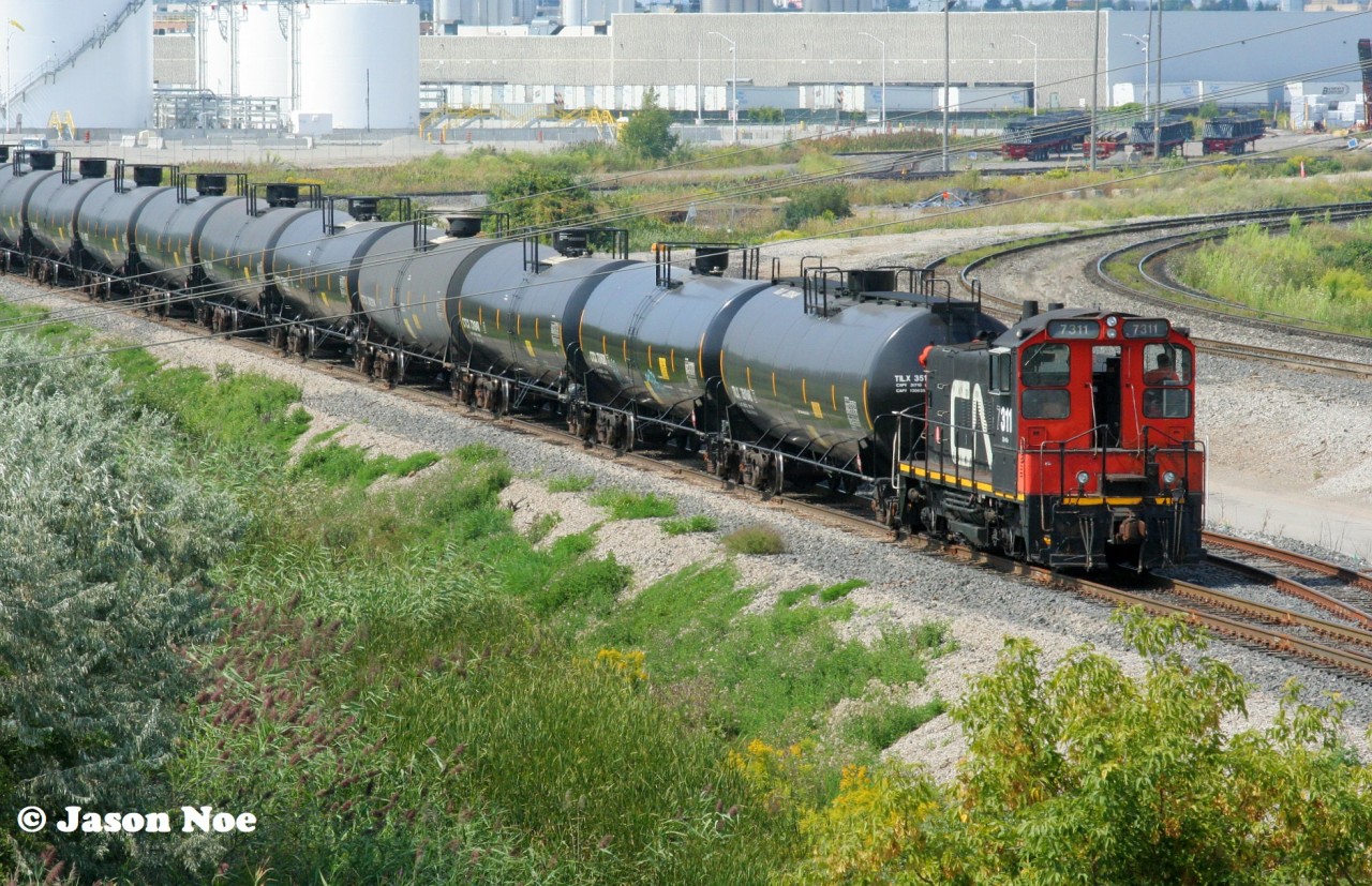 I would be lying if I said this one didn’t catch me by surprise. During my one visit this past August to the Highway #7 bridge that spans CN’s bustling MacMillan Yard in Vaughn, Ontario I spotted a CN switcher off in the distance deep within the yard. In 2023, CN had opened a new Cargoflo fuel transloading facility within the yard as well as constructed several storage tankers on site to store the fuel when unloaded. Situated off the W100 lead in the yard, CN had also built five tracks for this transload operation. Not long after my arrival on the bridge, the SW1200RSm came to life and began switching this facility. It’s been decades since I’ve photographed a CN switcher at MacMillan Yard, let alone working, so this was beyond cool to see in 2024. 

Here, CN SW1200RSm 7311 is viewed with a lengthy cut of tankers near the Highway #7 overpass with the fuel transloading facility in the background. Reportedly, both CN 7304 and 7311 are owned by a company under a CN subsidiary. However, only 7311 is the last remaining CN SW1200RSm on the current roster with its maintenance performed by CN at the MacMillan Yard Locomotive Reliability Center.