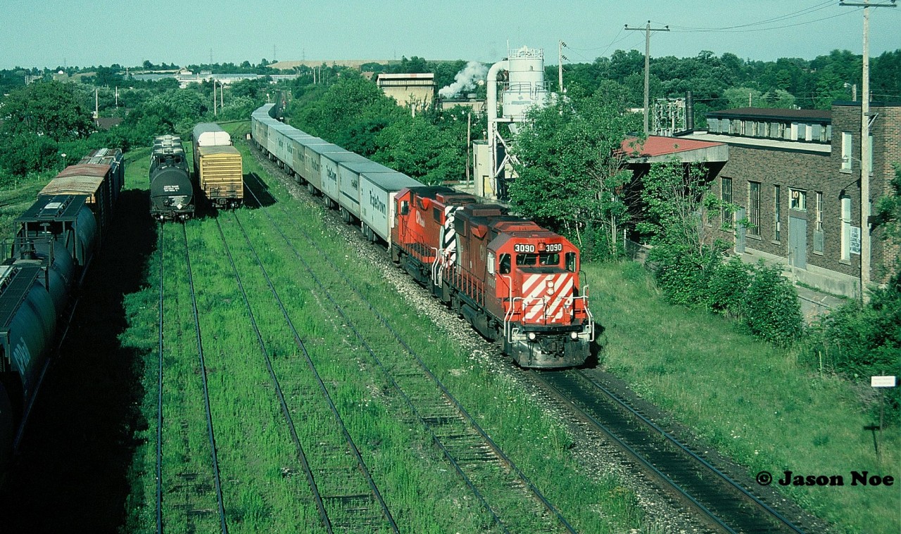 During a summer evening, the westbound CP Road Railer train 529 is viewed passing the east end of the yard in Galt, Ontario as it heads to London with a pair of multi-mark GP38-2’s. The tail-end of the train has just cleared the west siding switch Killean in the background.