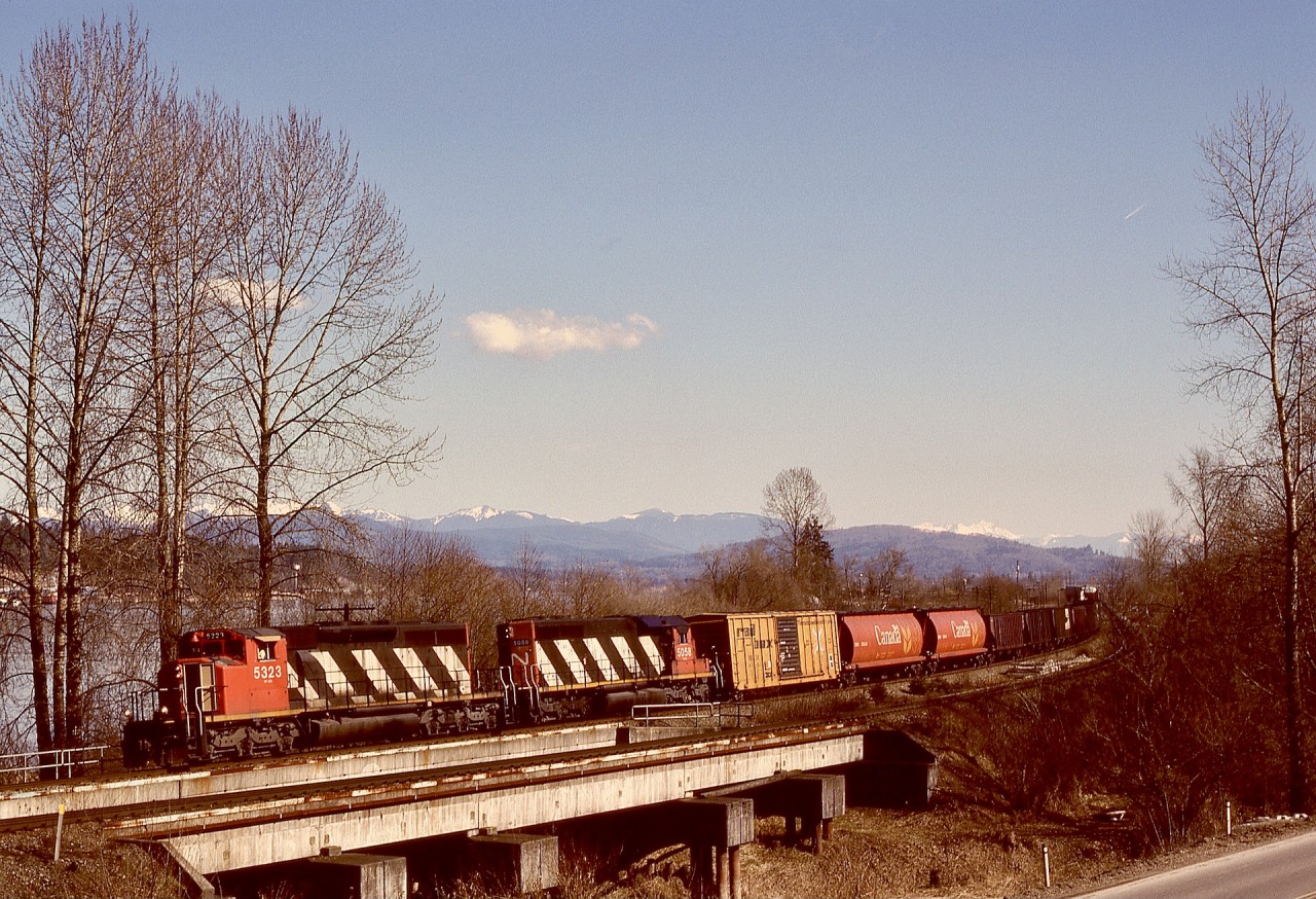 In following the Fraser River westward from Glen Valley, CN’s Yale subdivision squeezes by a bluff before reaching Fort Langley, and right at the beginning of that constriction it crosses West Creek at mileage 100.8.  On Sunday 1991-03-17, a westward mixed freight with CN 5323 + 5058 utilized the North Track at 1313 PST.

The silver-coloured water tower on the far bank of the Fraser River marks Whonnock on CP’s Cascade sub.