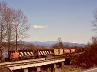 In following the Fraser River westward from Glen Valley, CN’s Yale subdivision squeezes by a bluff before reaching Fort Langley, and right at the beginning of that constriction it crosses West Creek at mileage 100.8.  On Sunday 1991-03-17, a westward mixed freight with CN 5323 + 5058 utilized the North Track at 1313 PST.

<p>The silver-coloured water tower on the far bank of the Fraser River marks Whonnock on CP’s Cascade sub.