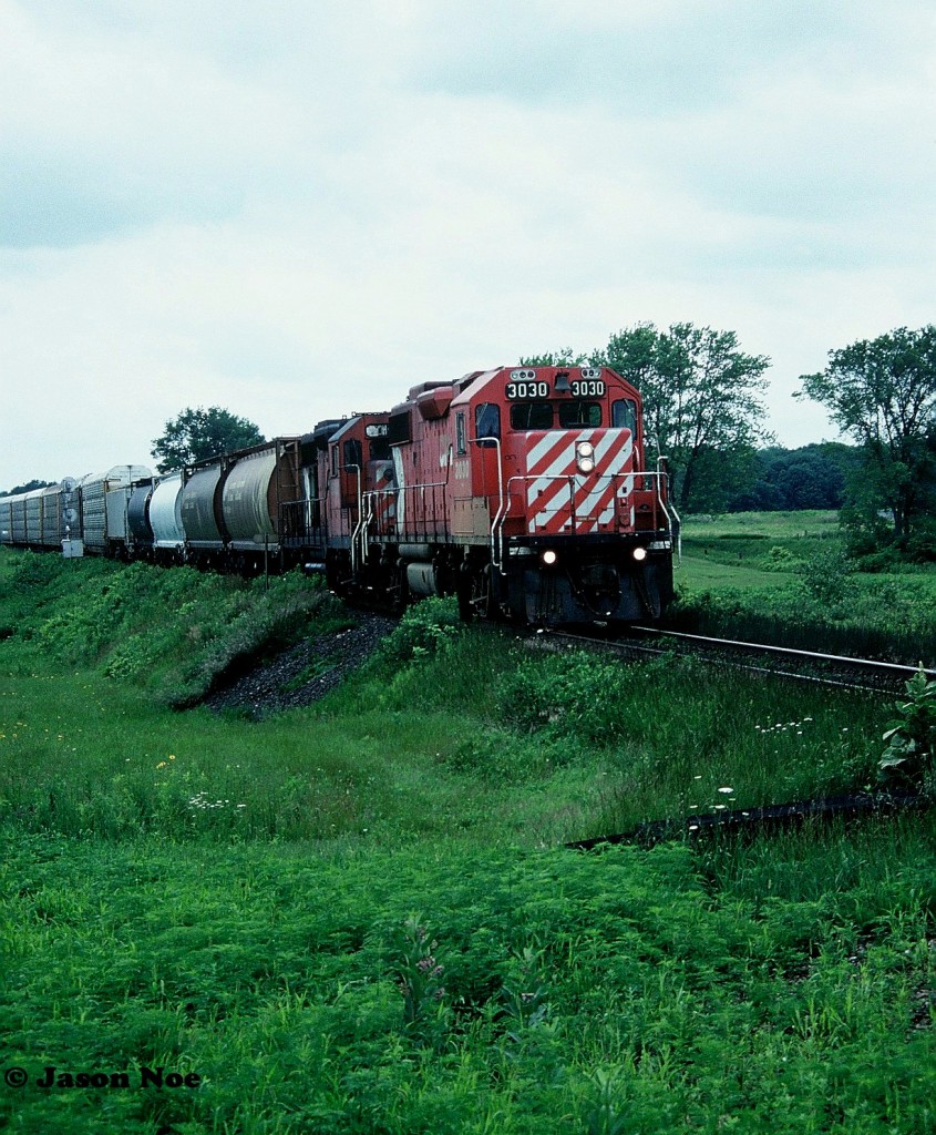 An eastbound CP train is viewed approaching the Komoka (Melrose) diamond crossing on the Windsor Subdivision, with the CN Strathroy Subdivision, situated west of London, Ontario. The consist included GP38-2 3030 and GP35 5012. At the time, the entire fleet of CP GP35’s was assigned to Western Canada and one straying this far east was very un-common. In August 1996 GP35 5012 was released from CP’s Weston Shops after conversion to control cab-daughter slug 1125. It was mated with GP38-2 ‘mother’ 3035 and assigned to Winnipeg. It was ultimately retired in October 2012.