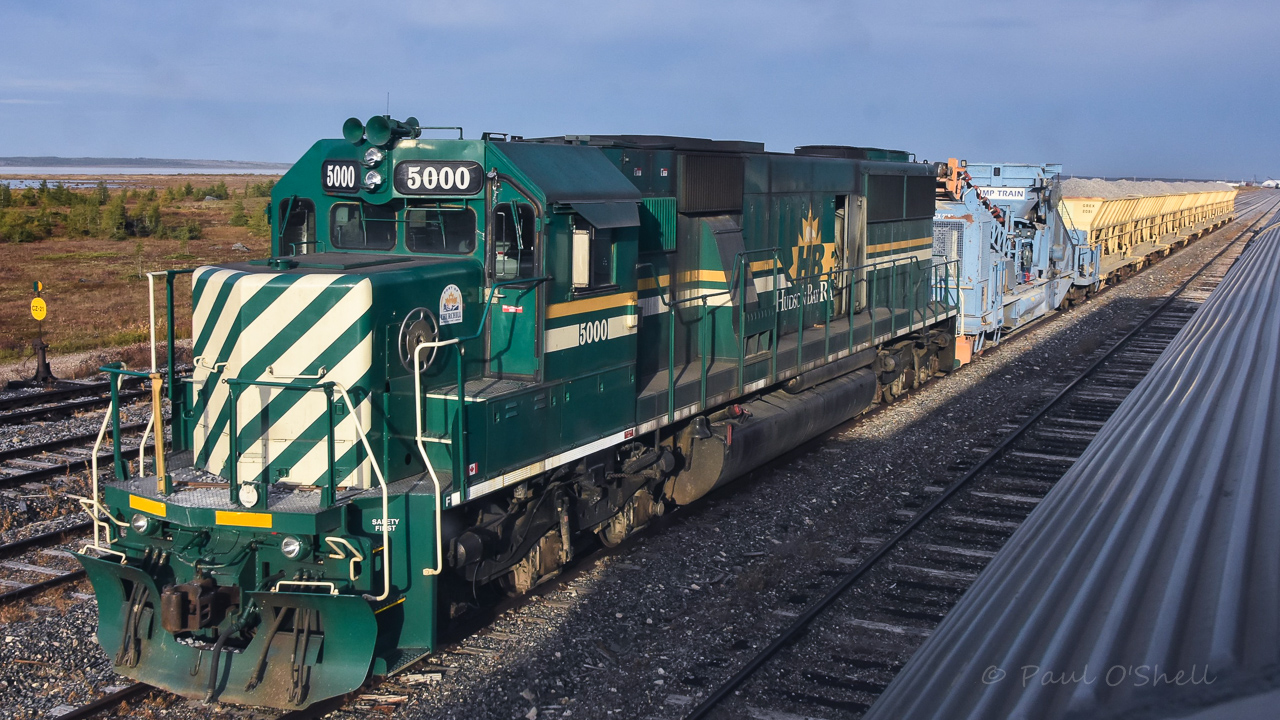 Green Power  
As VIA 693 The Hudson Bay backs its way slowly to the station track in Churchill, HBRY 5000 stands ready to move out with a loaded GREX ballast unloading Dump Train from the HBRY Yard at Mile 509.8 Herchmer Sub. on what is shaping up to be a gorgeous September 19, 2024 da in this wonderful remote location. 
Another of my pics graciously edited by Benny :-)