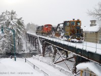 Just off Muskoka Road is a neat little bridge in the town of Bracebridge, and CN spreader "W908" is heading north on the Newmarket Subdivision clearing some of the 3 feet of snow that fell in the days prior. With a sudden and severe cold snap having come in January this meant warmer waters dumped all their moisture as snow over days with local accumulations topping out at 50 cm per day. CN would drag this spreader out of the MOW track in Stratford to bring it to Toronto and run it north on the Bala and Newmarket subs. The building in the background screams cold with large icicles dangling from the roofline. Now owned by the Town of Bracebridge this building was Bird Woolen Mill built in 1918. <a href=https://engagebracebridge.ca/3ecclestone/news_feed/property-background target=_blank>Source</a>.