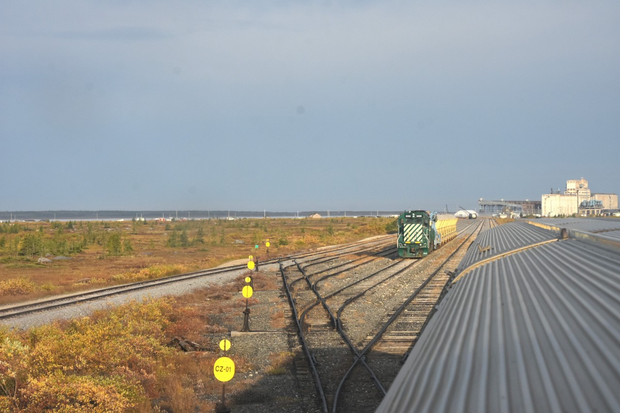 Ladies & Gentlemen, Welcome to Churchill  
VIA 693 The Hudson Bay makes its 5 mph reverse movement from HBR Churchill South, through the recently rehab'd Churchill yard, towards the stately station in Churchill, MB. 
The day is starting out great with sunshine and just a bit of hazy sky. 
Not much in the yard this early September morning except a loaded GREX Dump Train, a few tank cars, and a single bulkhead flat loaded with rolls of Geotextile fabric for laying down during roadbed rehabilitation. 
I'm just 10 minutes away from detraining and starting a full day of exploring the town and HBRY infrastructure once again. :-)