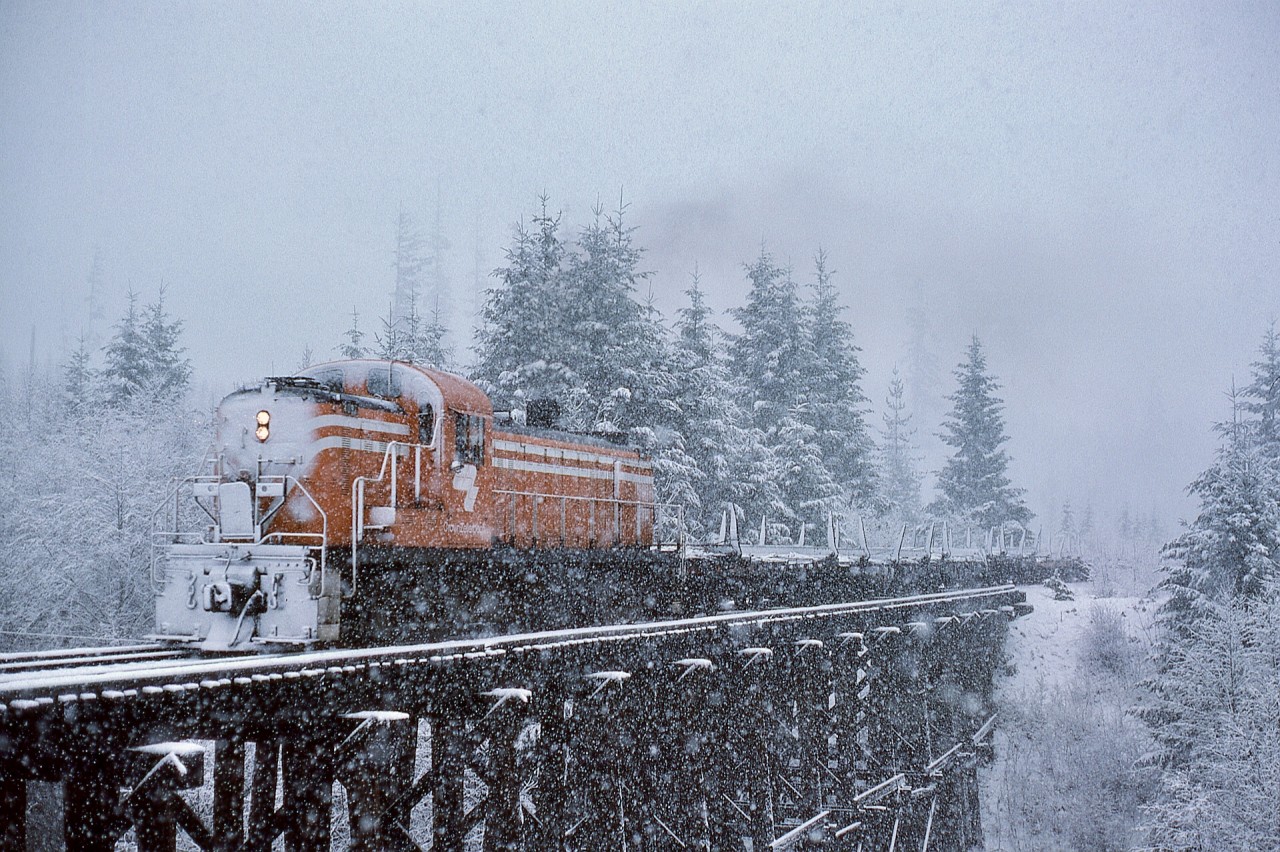 Westward from Ladysmith, Crown Zellerbach’s log-haul railway ascended to Nanaimo Lakes, so what was rain down at Ladysmith often fell as snow farther up.  That was the case on Wednesday 1976-02-25 as ALCo RS-3 4097 (ex D&H 4117) hauled empty log flats across a timber trestle over Boulder Creek at mileage 12.2 enroute a reload up at Nanaimo Lakes.

An unusual operating arrangement is shown in this photo.  Crown Zellerbach and other tidewater-to-inland logging railways almost always ran their steam locomotives with the smoke box oriented upgrade for best firebox crownsheet coverage, and that front-to-the-woods plan was carried through into the diesel era.  However, to counter increased wheel wear on one side of 4097, it was run short-nose-upgrade for several months, and I was fortunate to catch several examples of that.

Wishing you all a Merry Christmas and Happy Holidays.