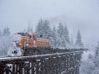 Westward from Ladysmith, Crown Zellerbach’s log-haul railway ascended to Nanaimo Lakes, so what was rain down at Ladysmith often fell as snow farther up.  That was the case on Wednesday 1976-02-25 as ALCo RS-3 4097 (ex D&H 4117) hauled empty log flats across a timber trestle over Boulder Creek at mileage 12.2 enroute a reload up at Nanaimo Lakes.

<p>An unusual operating arrangement is shown in this photo.  Crown Zellerbach and other tidewater-to-inland logging railways almost always ran their steam locomotives with the smoke box oriented upgrade for best firebox crownsheet coverage, and that front-to-the-woods plan was carried through into the diesel era.  However, to counter increased wheel wear on one side of 4097, it was run short-nose-upgrade for several months, and I was fortunate to catch several examples of that.

<p>Wishing you all a Merry Christmas and Happy Holidays.