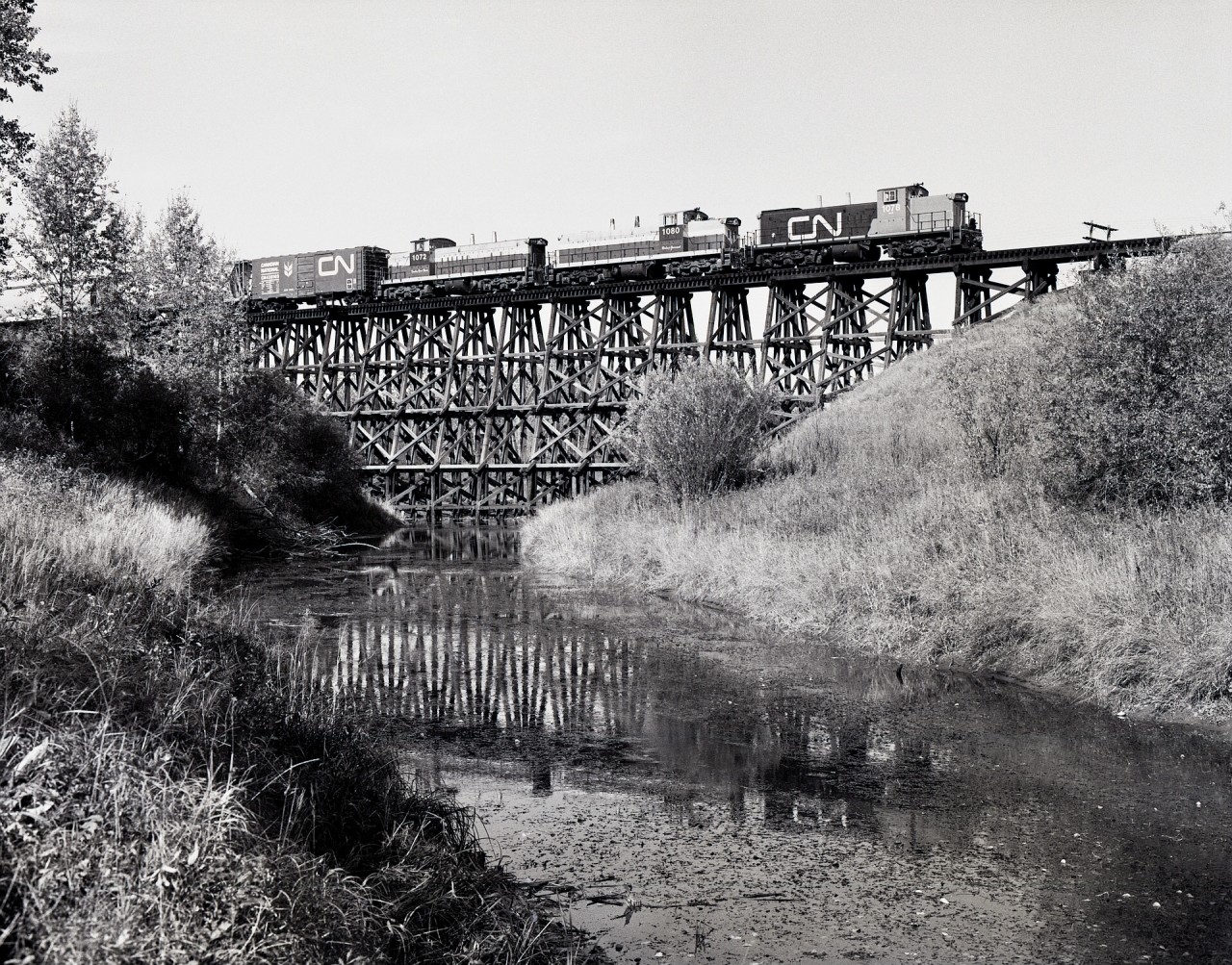 Heading for Edmonton’s ex NAR Dunvegan Yards on its second day of travel from Waterways, a Mixed Extra South with CN 1078 + 1080 (ex NAR 303) + 1072 (ex NAR 311, nee CN 1072) is crossing the timber trestle over the Sturgeon River at mileage 9.7 on Wednesday 1981-09-30 at 1232 MDT.

Until the end of 1980, this was Northern Alberta Railways’ Edmonton sub., logically renamed to the CN Westlock sub. when they took over.  Somewhat to my surprise, aside from renumbering NAR locomotives to the CN plan, the NAR diamond symbol and name and unit names (Bishop Grouard and Twelve Foot Davis) were left intact in the first CN year.