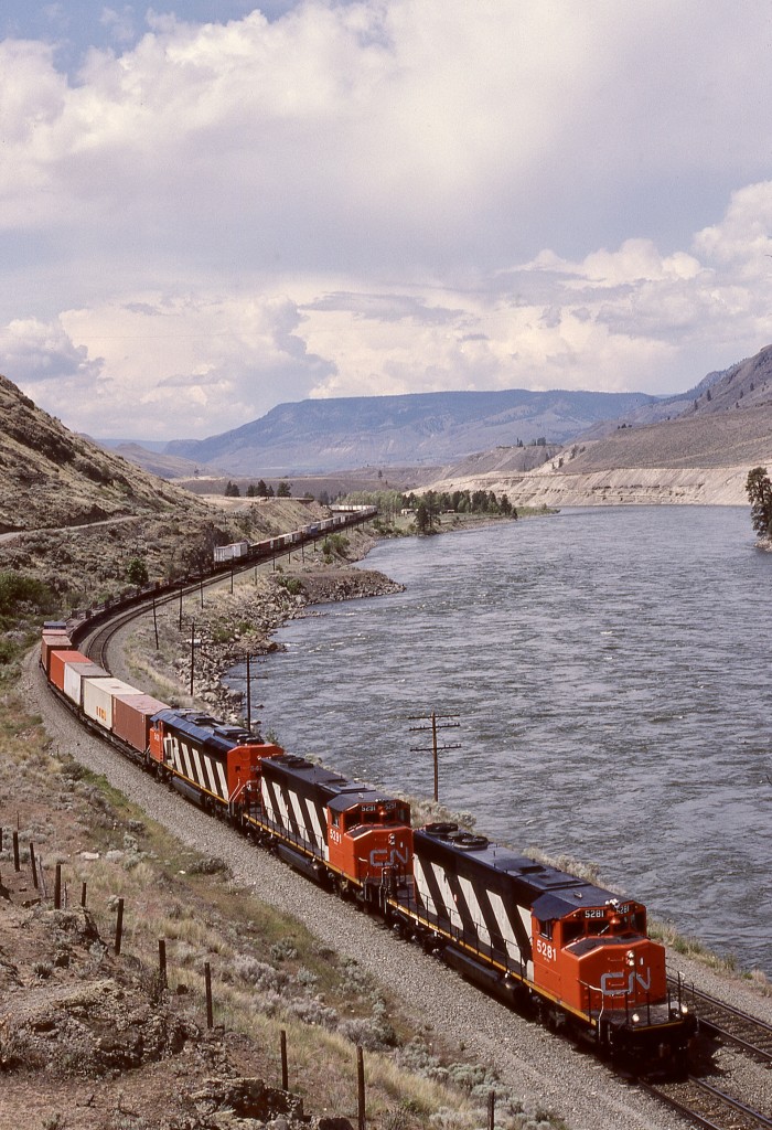 Running alongside the Thompson River between Ashcroft and Savona and enroute next crew change at Kamloops on Thursday 1987-05-21, an eastward intermodal freight powered by CN 5281 + 5291 + 5439 is at mileage 31.8 and just about at the east end of the siding at Walhachin.