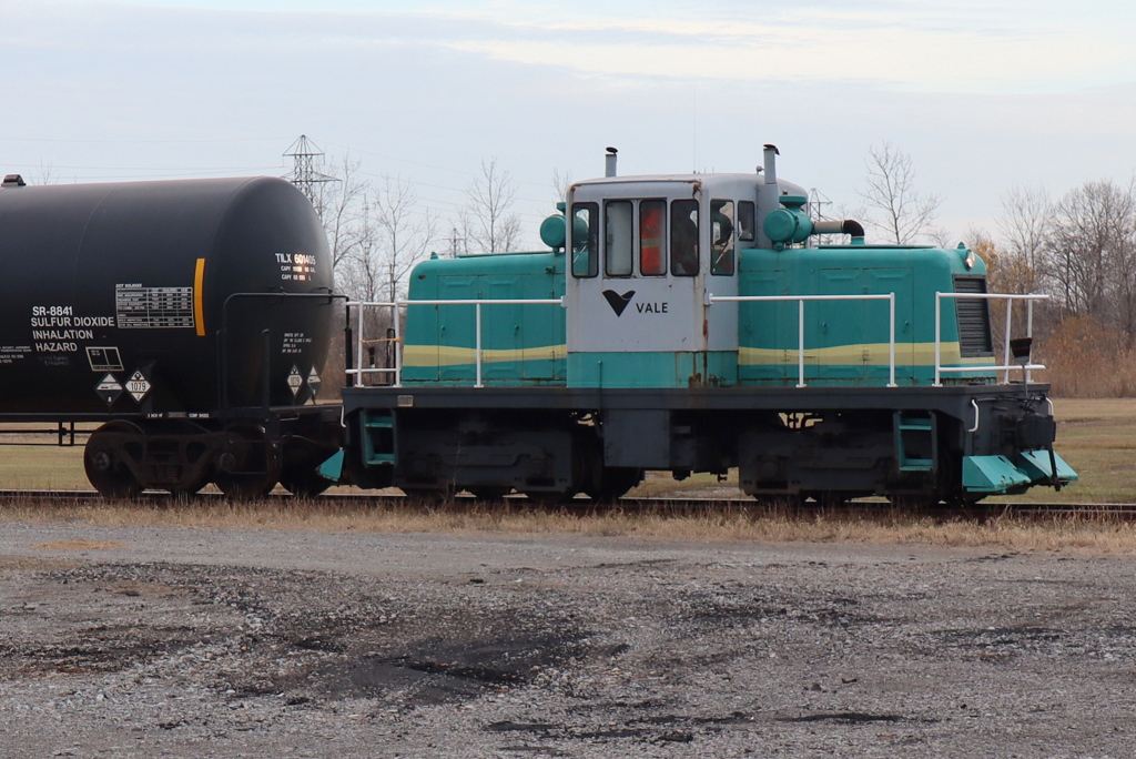 This often-illusive critter is returning and deeper into Vale’s compound with a single tank car just picked up from the inbound track previously set off by CN.  When passing by on Durham St, an inbound car was evident, but something seemed different regarding the critter’s customary parking spot.  By the time that I had bypassed construction and traffic to reach the outside parking lot, the crew had already spotted an empty car on the outbound track. The 2-person crew assisting the move conveniently scooted about in an EZGO (22) 4-wheeler. The Trackside Guide describes this VALE critter as GE, serial 31716, 2/1953 and 45T … acquired new, ballasted to 50T. 
Is this colourful critter one and the same as the INCO yellow critter captured by Paul Chapman 12/18/1980 and recently submitted by / from Dean Brown’s collection? Unique covers have been added in the interim, as a safety precaution most likely, to prevent riding on the footboards per se.