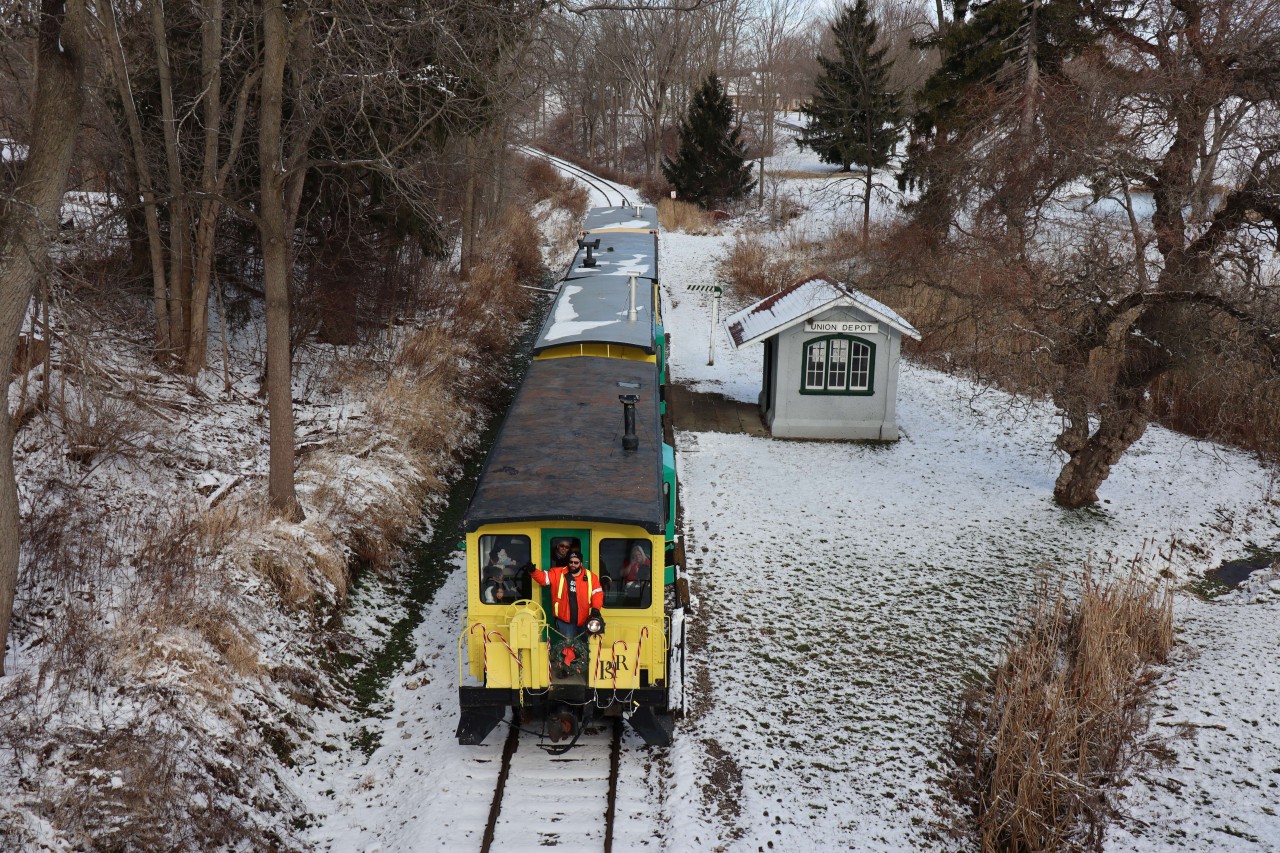 PSTR Santa Treats Train (1st ride of 3 this day) is returning to Port Stanley from Whyte’s Park.  The train consisting of three cars 66, 62 and 63 plus the Little Red Caboose 602, filled with excited children and parents, is alongside Union Depot (previously the North Pole). Port Stanley Terminal Rail’s L1 Stanley (although perhaps not visible) is pushing its Christmas train downgrade through the Kettle Creek Valley on the historic tracks of Port Stanley and London Railway. Santa Claus hopped on and off the train, this year, when passing through the storage yard located a short distance from the PSTR station.  The timely skim of snow overnight adds to the scene and festive occasion. 
This photo may qualify for the Time Machine in conjunction with a photo by Greg Roach ID: 21596 taken at Union (49 years previous) on a UCRS Excursion from Toronto to Port Stanley.