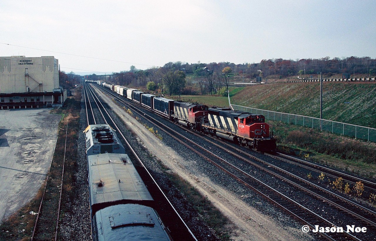 CN SD40-2(W) 5354 and GP40-2L(W) 9631 are viewed bringing an eastbound into Aldershot Yard on the Oakville Subdivision. To the left, VIA Rail train 73 is departing the Aldershot station for the Dundas Subdivision with 6457 west.