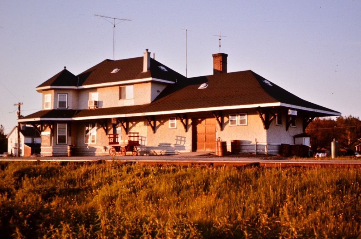 Beautiful downtown Oba, ON  
Another view of the CN Oba, ON station from early July 1976. 
The platform and surrounding grounds are littered with common place items (at the time) seen at stations throughout the north. 45-gallon drums of gasoline, propane tanks and oil (kerosene) drums for the station agent and work crews in the yard. A freshly painted baggage cart rests in front of the station waiting on its next load of arriving or departing cargo. 
Lots of antennas on the roof for tv and radio communications.