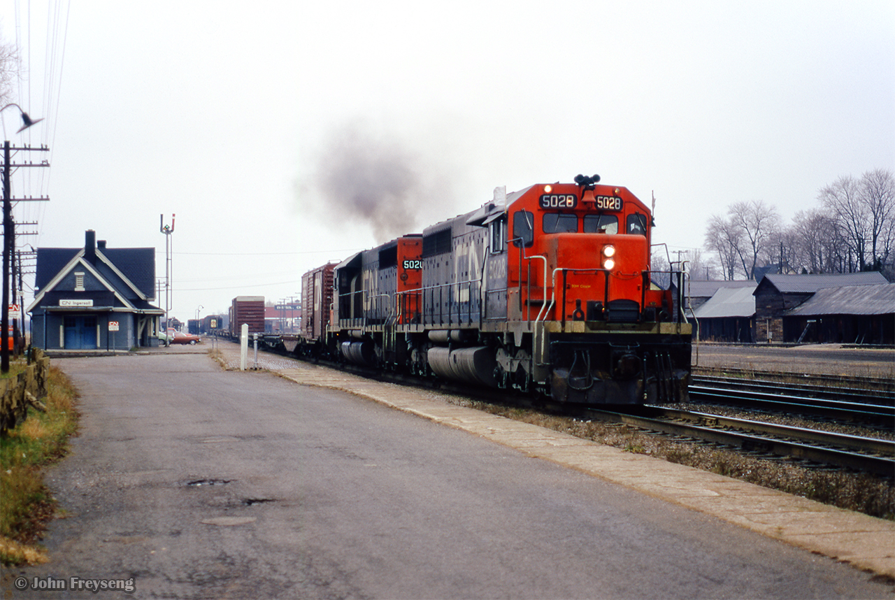 After setting off a cut of hoppers (seen just above the red car's hood,) CN Extra 5028 east thunders past the station at Ingersoll for points east.  Though hard to see, an SW1200RS is in the yard beyond the train order signal.
Scan and editing by Jacob Patterson.